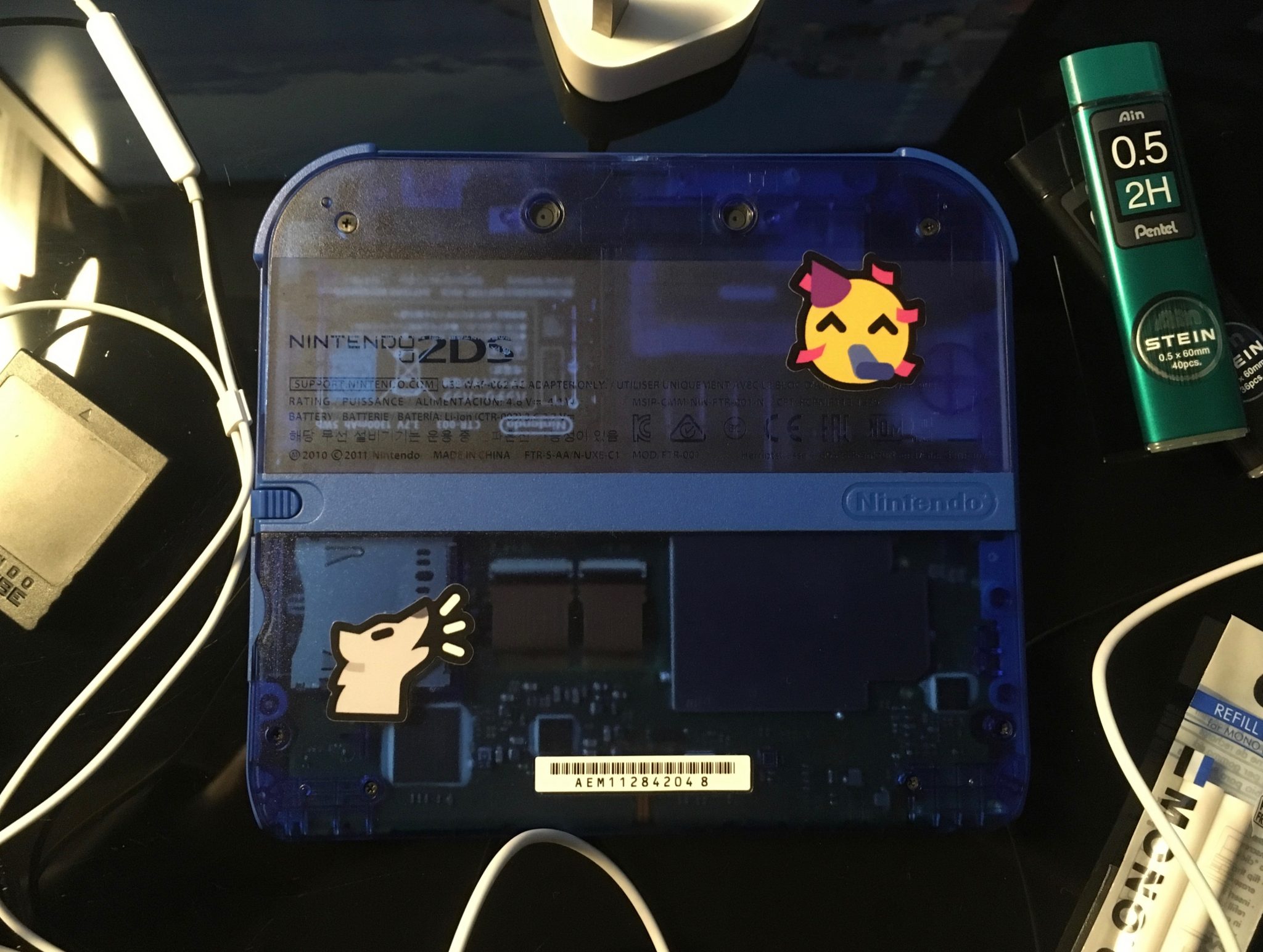 The back of a transparent blue Nintendo 2DS face down on a desk. It has a party face sticker on the top right and an awoo sticker on the bottom left. It is surrounded by different objects - white headphone cable, Nintendo GameCube black memory card and mechanical pencil and eraser refills.