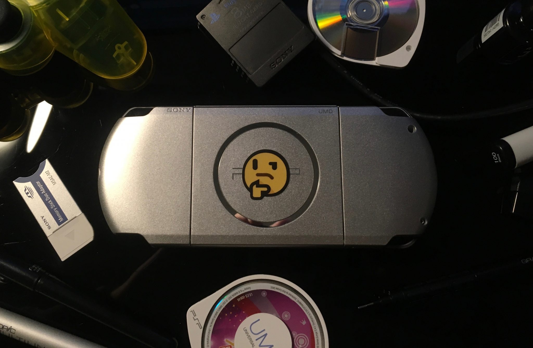 A silver PSP face down on a black glass desk. In the center of the back of a console is a thinking emoji sticker. Surrounding it are different scattered objects - a transparent yellow PlayStation 2 controller, UMDs, a PS2 memory card, black nail varnish, a Memory Stick Duo to Memory Stick adaptor, and various kinds of pens and pencils.