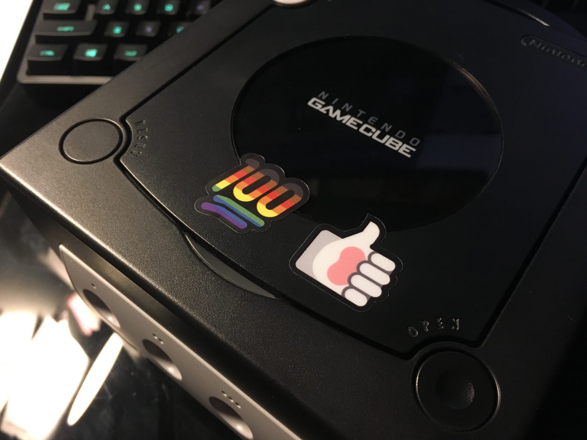 A pride 100 and a paw thumbs up sticker stuck on the lower part of the disc tray cover on a black Nintendo GameCube.