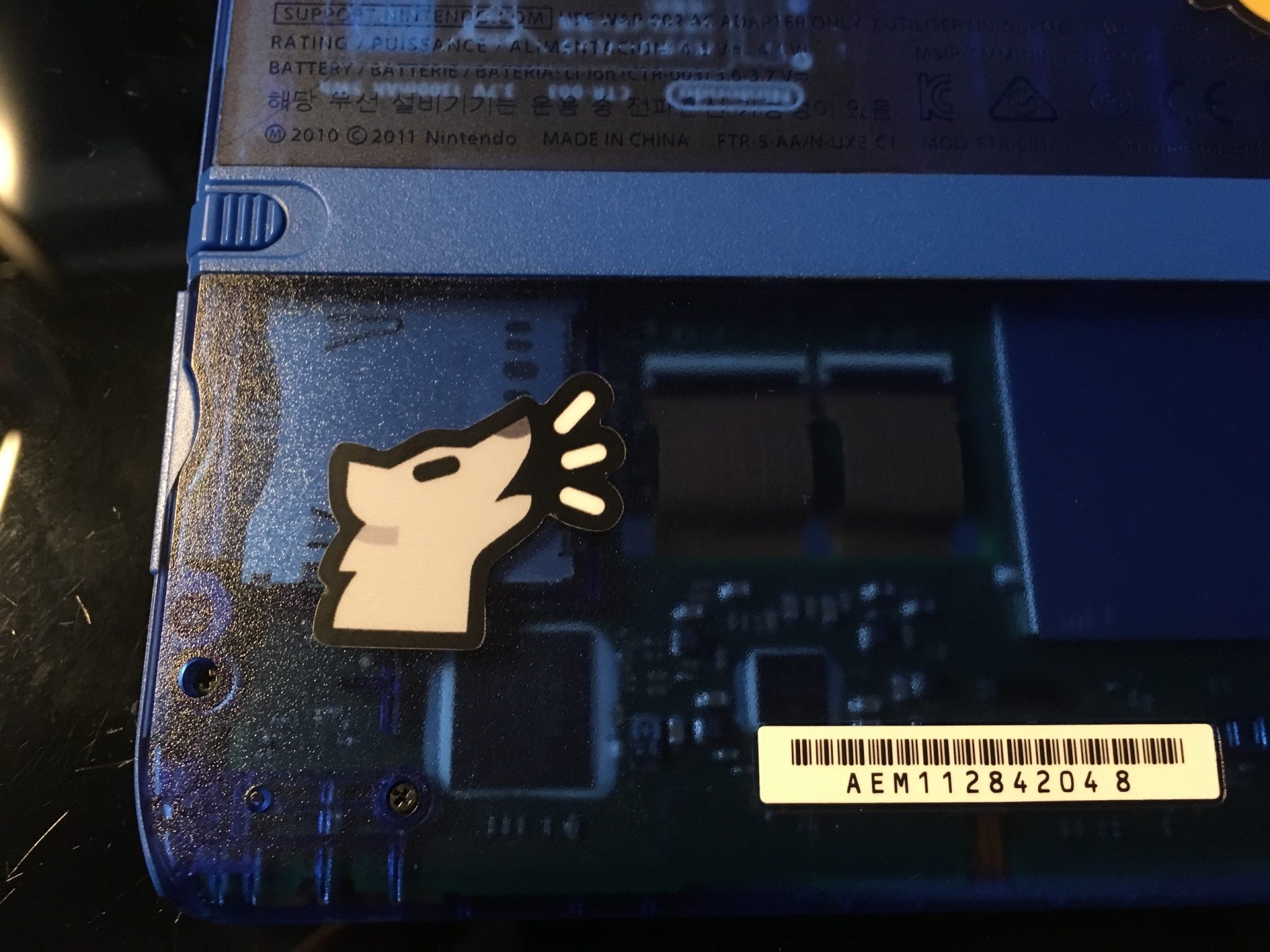 A detailed shot of an Awoo sticker on the bottom right of the back of a transparent blue Nintendo 2DS. With a semi-warm lamp shining on it from the left hand side.